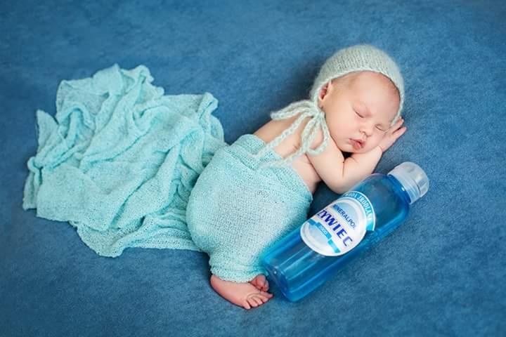 Śpiące niemowlę w błękitnym stroju i czapeczce leży na błękitnym tle obok butelki wody mineralnej Żywiec Zdrój.