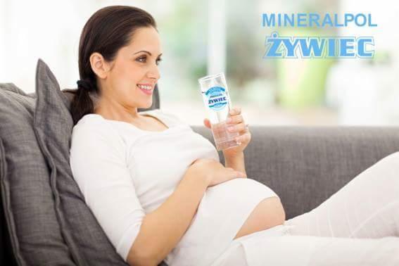 Uśmiechnięta kobieta w ciąży pije wodę mineralną Żywiec, siedząc na szarej sofie.