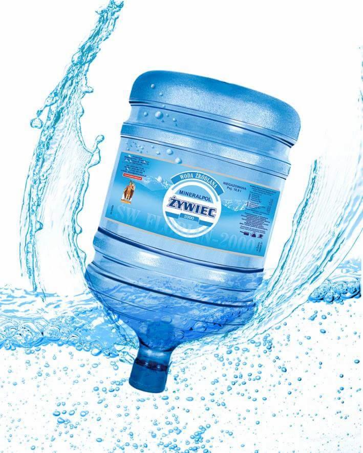 Duży, przezroczysty baniak wody Żywiec Zdrój otoczony rozpryskującą się wodą na białym tle.