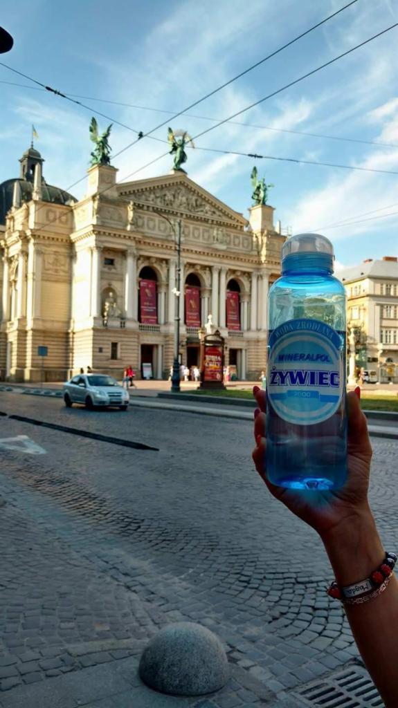 Błękitna butelka wody Żywiec w dłoni na tle Opery Lwowskiej, widok z brukowanej ulicy.