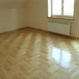 Pusty pokój na poddaszu z parkietem ułożonym we wzór jodełki, biały grzejnik pod oknem, ściany przygotowane do malowania.