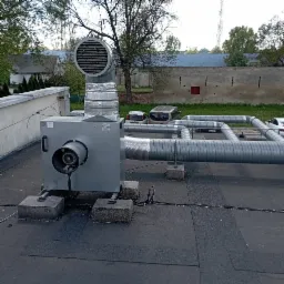 Szara jednostka centralna wentylacji mechanicznej z rurami wentylacyjnymi na dachu budynku, widoczne betonowe podstawy i przewody elektryczne.