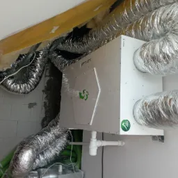 Centrala wentylacyjna z rekuperacją SmartVent zamontowana w pomieszczeniu, widoczne aluminiowe kanały wentylacyjne, ściany z białych bloczków oraz żółta izolacja stropu.