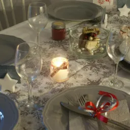 Uroczysty stół wigilijny z szarymi talerzami, kieliszkami, świecami w słoikach z kokardkami, dekoracjami w szklanych naczyniach i kartami z imionami gości na tle obrusu w śnieżynki.