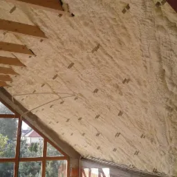 Poddasze ocieplone pianką poliuretanową, widoczne krokwie i okno w murze z czerwonej cegły, szare łączniki w pianie.