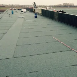 Prace na dachu hali: dekarz zgrzewa papę termozgrzewalną, widoczne butle gazowe, narzędzia i częściowo wykonana izolacja.