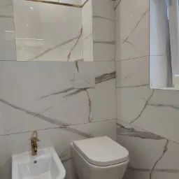 Nowoczesna łazienka z białymi płytkami imitującymi marmur z złotymi akcentami, wiszącą toaletą i bidetem z baterią w kolorze złota.