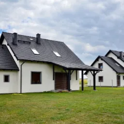 Nowoczesne domy jednorodzinne z ciemnymi dachami i jasną elewacją, widoczne okna z roletami zewnętrznymi, brama garażowa z roletą, trawnik przed budynkami, pochmurne niebo.