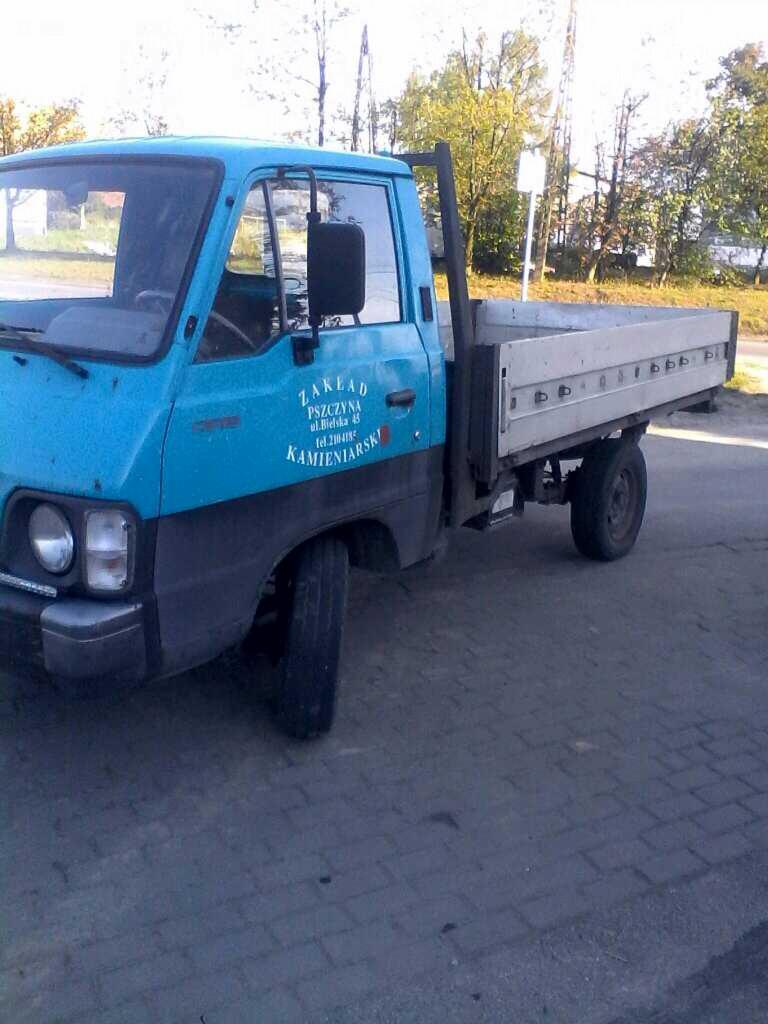 Niebieski, lekko zardzewiały samochód dostawczy z otwartą skrzynią ładunkową, zaparkowany na brukowanej powierzchni. Na drzwiach widoczny napis 'Zakład Kamieniarski Pszczyna'.
