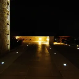 Nocne oświetlenie ścieżki z kamiennej kostki prowadzącej do balustrady z widokiem na odległe światła miasta, z podświetloną kamienną ścianą po lewej stronie.