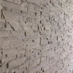 Ściana wykończona dekoracyjną, szarą okładziną kamienną o nieregularnej fakturze i poziomym układzie, oświetlona górnym światłem.