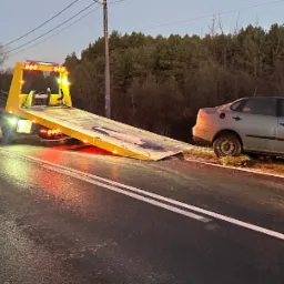Żółta laweta z włączonymi światłami awaryjnymi na drodze, przygotowuje się do załadunku uszkodzonego, srebrnego samochodu osobowego z otwartą maską.