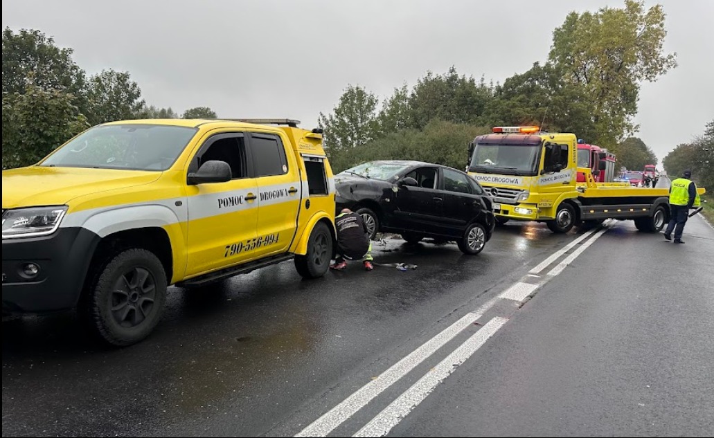 Dwa żółte pojazdy pomocy drogowej zabezpieczają miejsce kolizji czarnego samochodu osobowego na mokrej, asfaltowej drodze. Na pierwszym planie pracownik w stroju roboczym przygląda się uszkodzeniom...