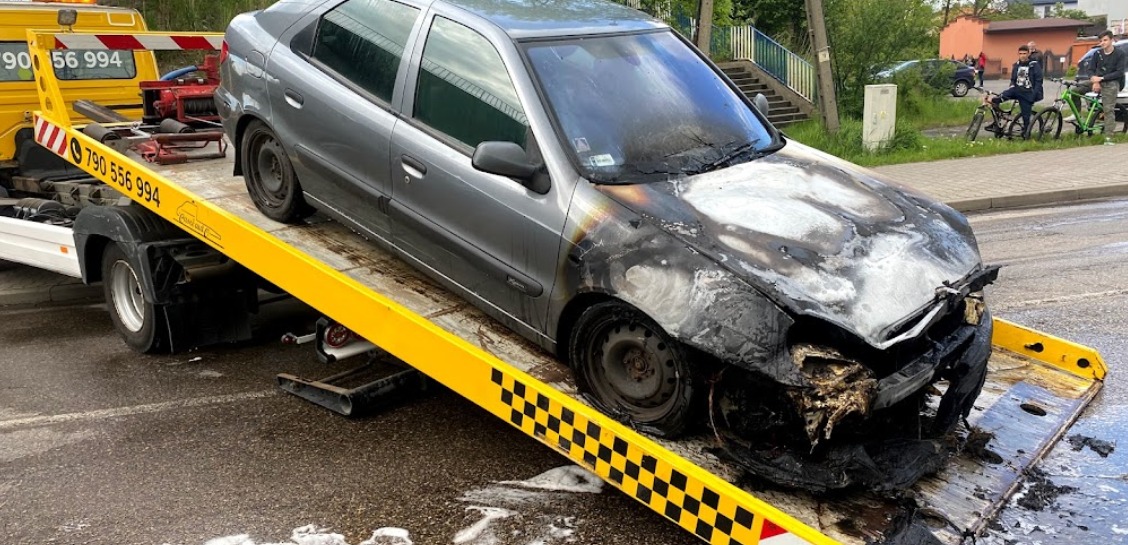 Uszkodzony w wyniku pożaru szary samochód osobowy na żółtej lawecie z czarno-białą szachownicą, widoczne zwęglone elementy karoserii, tło miejskie z pieszymi i rowerzystami.