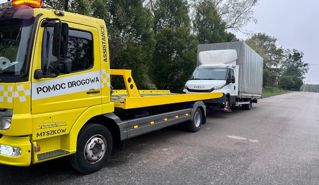 Żółta laweta z napisem 'Pomoc Drogowa' holuje biały samochód dostawczy Iveco na szarej, mokrej drodze, widok z boku.