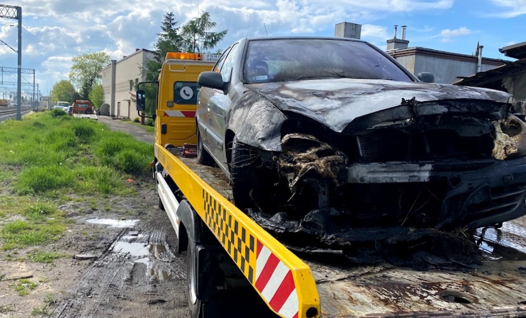 Uszkodzony, czarny samochód osobowy z widocznymi zniszczeniami przodu, załadowany na żółtą lawetę z pomarańczowym światłem ostrzegawczym, stojącą na błotnistym terenie w pobliżu torów kolejowych.