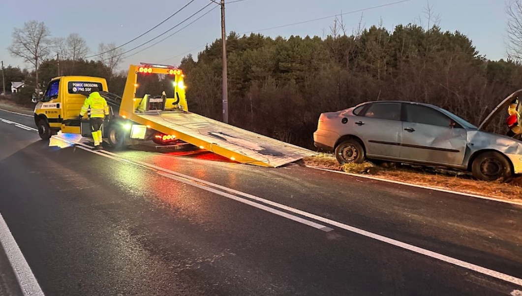 Żółta laweta z włączonymi światłami awaryjnymi na drodze, przygotowuje się do załadunku uszkodzonego, srebrnego samochodu osobowego z otwartą maską.