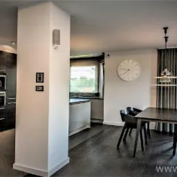 Nowoczesne wnętrze: widok z jadalni na kuchnię, ciemne drewniane podłogi, białe ściany, minimalistyczny stół z krzesłami, designerska lampa wisząca i wbudowany ekspres do kawy.