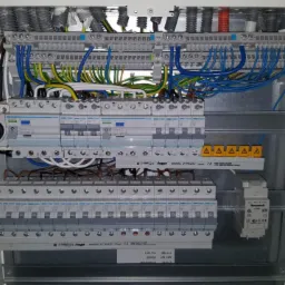 Otwarta skrzynka rozdzielcza z licznymi bezpiecznikami, przewodami w różnych kolorach i modułami marki Hager oraz Honeywell, zamontowana w białej obudowie.