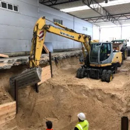 Żółta koparka New Holland w trakcie prac ziemnych przy zabezpieczonym wykopie, widoczni pracownicy w odblaskowych ubraniach i kaskach, w tle budynek przemysłowy z widoczną konstrukcją dachu.