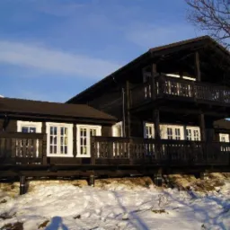 Dwupiętrowy, ciemny dom z drewna z balkonami i białymi oknami, posadowiony na śnieżnym zboczu z widokiem na zaśnieżoną górę w tle.