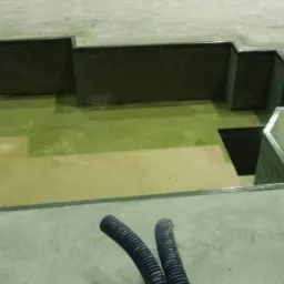 Wykop z betonowymi ścianami i dnem, częściowo wypełniony wodą, widoczne dwa karbowane węże na pierwszym planie.