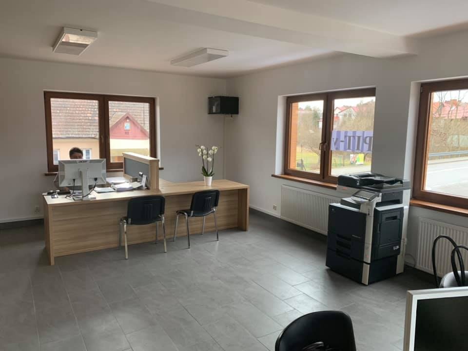 Jasne wnętrze biura z drewnianym biurkiem, dwoma monitorami, krzesłami, drukarką i kwiatami w wazonie, widok zza okna na budynki mieszkalne.