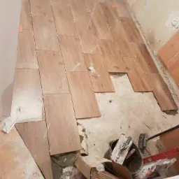 Ułożone płytki podłogowe imitujące drewno, widoczne plastikowe krzyżyki dystansowe, fragmenty nieukończonej podłogi, narzędzia glazurnicze w wiadrze i poziomica.