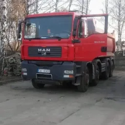 Czerwona ciężarówka MAN TGA 41.440 z otwartymi drzwiami kierowcy, zaparkowana na betonowym placu z drzewami w tle.