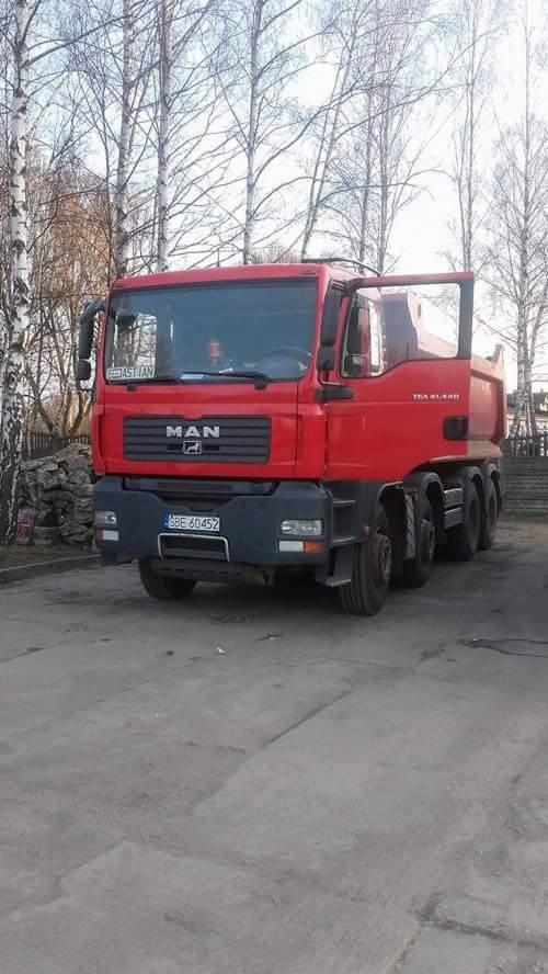 Czerwona ciężarówka MAN TGA 41.440 z otwartymi drzwiami kierowcy, zaparkowana na betonowym placu z drzewami w tle.