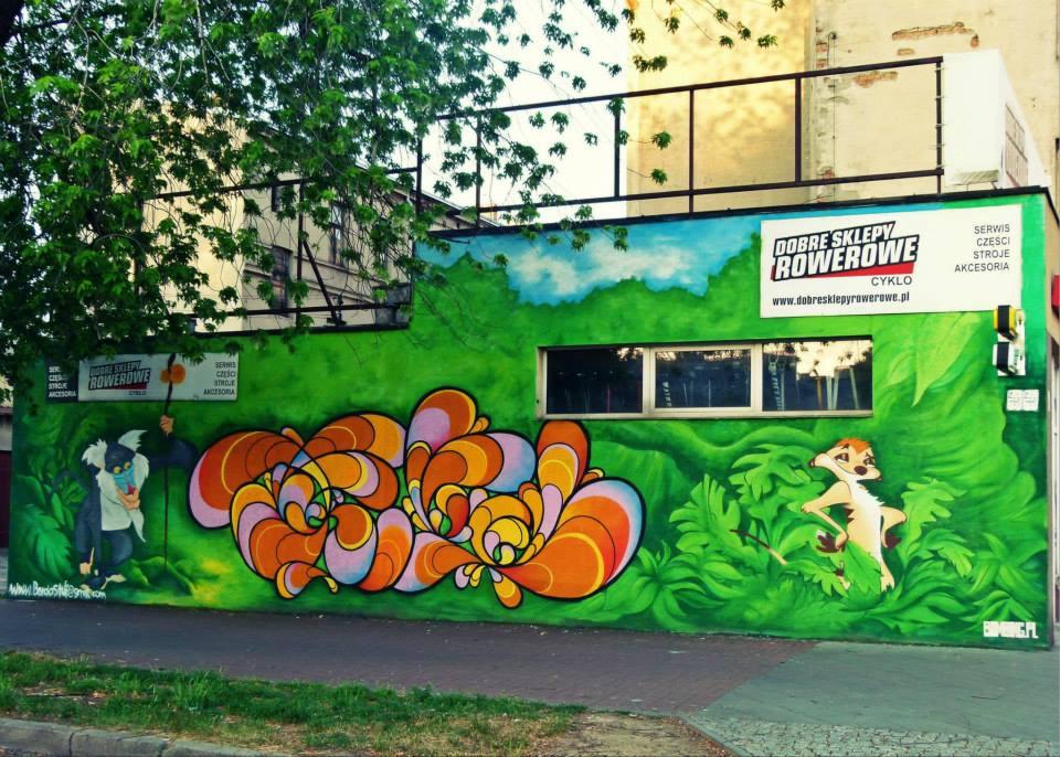 Zewnętrzna ściana budynku z kolorowym muralem przedstawiającym abstrakcyjne wzory, postacie zwierząt i logo sklepu rowerowego na tle zieleni i błękitnego nieba.