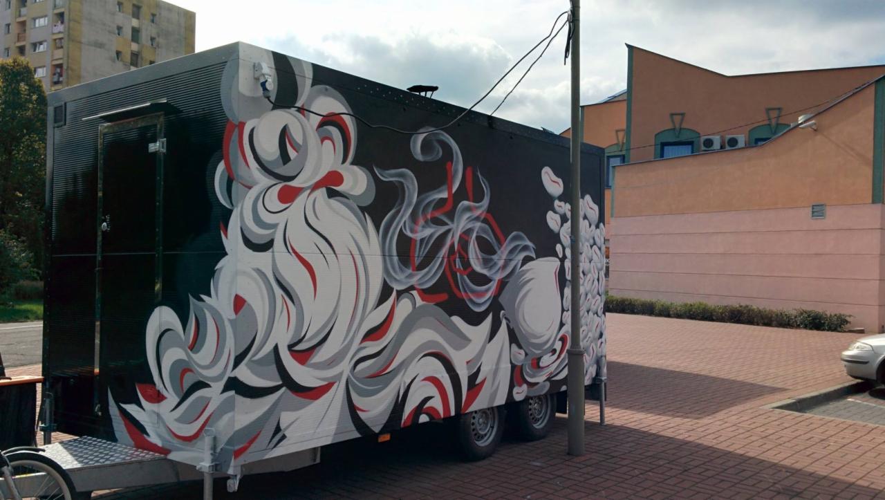 Czarna przyczepa z abstrakcyjnym, wielokolorowym malunkiem w odcieniach bieli, szarości i czerwieni, stojąca na brukowanej powierzchni, z budynkami w tle.