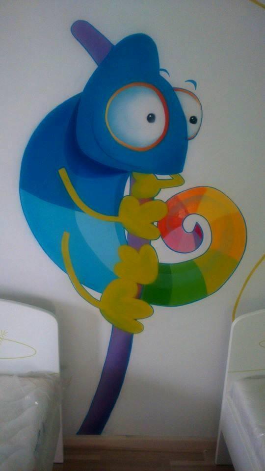 Kolorowy mural z wizerunkiem kameleona na ścianie w pokoju dziecięcym, widoczne łóżeczka dziecięce.