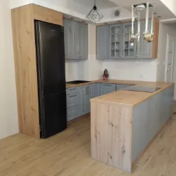 Nowoczesna kuchnia z szarymi szafkami, drewnianymi blatami i czarną lodówką w zabudowie; widoczne elementy oświetlenia sufitowego.