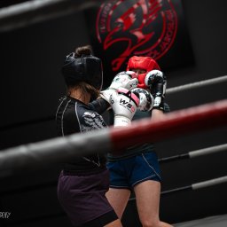 Photography by Denis Rubelt - Dwie osoby w kaskach i rękawicach bokserskich walczące na ringu, widoczne logo klubu sportowego w tle.