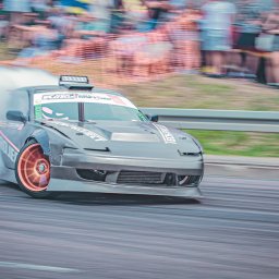 Photography by Denis Rubelt - Szary samochód do driftu w trakcie jazdy, z widocznymi śladami opon i dymem, na tle rozmytej publiczności i wozu strażackiego.