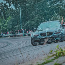 Photography by Denis Rubelt - Szary samochód BMW M3 driftuje na zakręcie toru, widzowie obserwują za barierką, widoczne ślady opon na asfalcie.