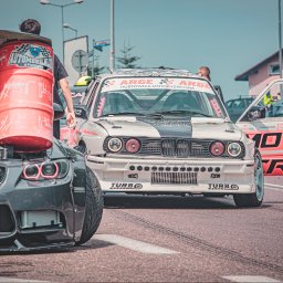 Photography by Denis Rubelt - Dwa samochody wyścigowe na torze, jeden z otwartą maską i beczką z logo Automobilklubu, drugi z otwartymi drzwiami i naklejkami sponsorów