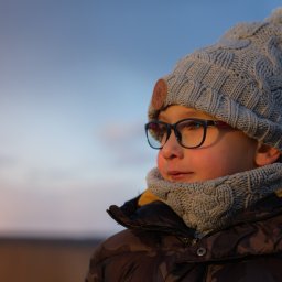 Photography by Denis Rubelt - Portret chłopca w okularach i zimowej czapce z pomponem, spoglądającego w bok na tle rozmytego nieba o zachodzie słońca.