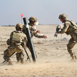 Photography by Denis Rubelt - Żołnierze w kamuflażu obsługują moździerz na piaszczystym poligonie, unoszący się pył, jeden żołnierz podaje amunicję.