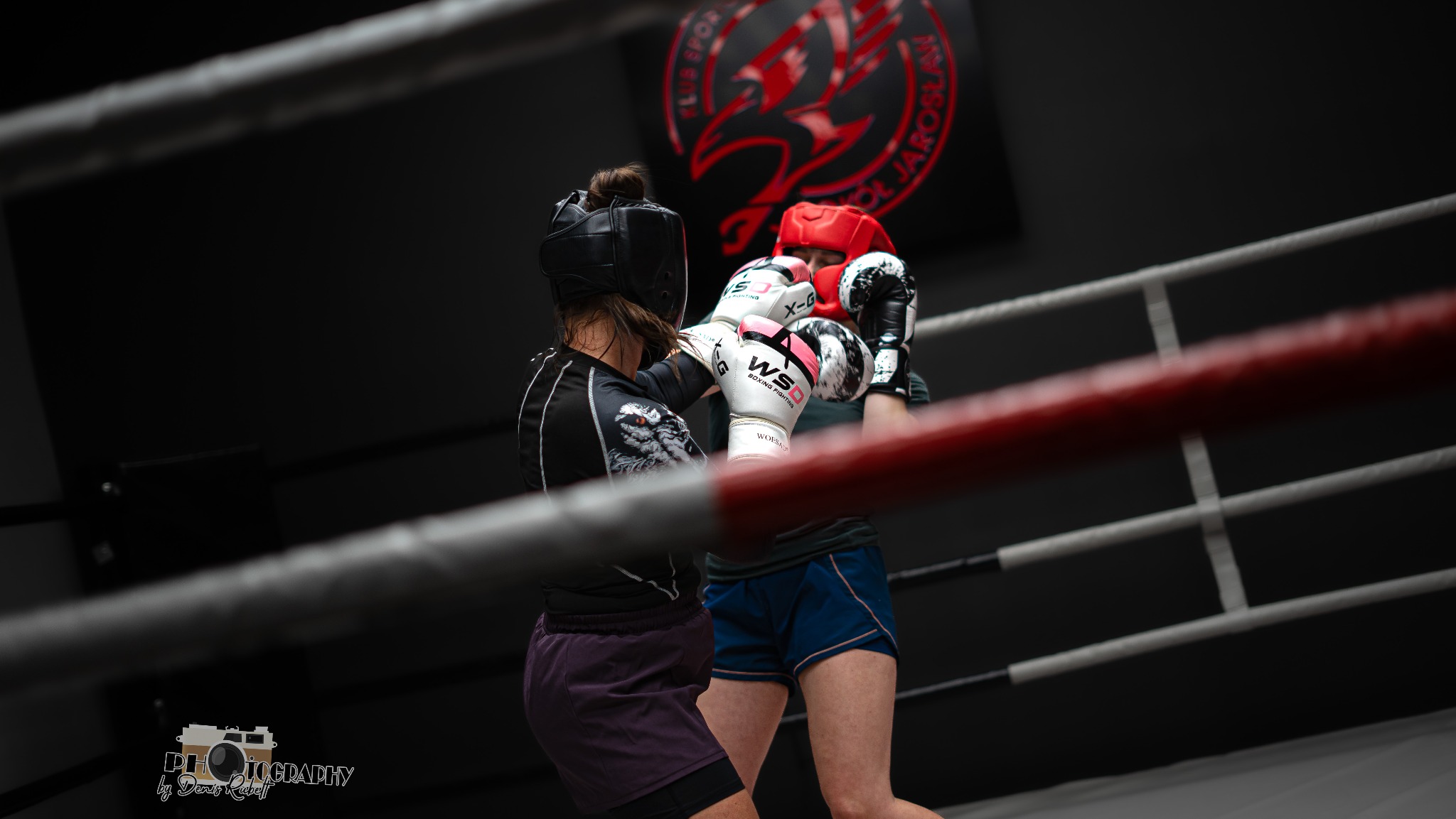 Dwie osoby w kaskach i rękawicach bokserskich walczące na ringu, widoczne logo klubu sportowego w tle.