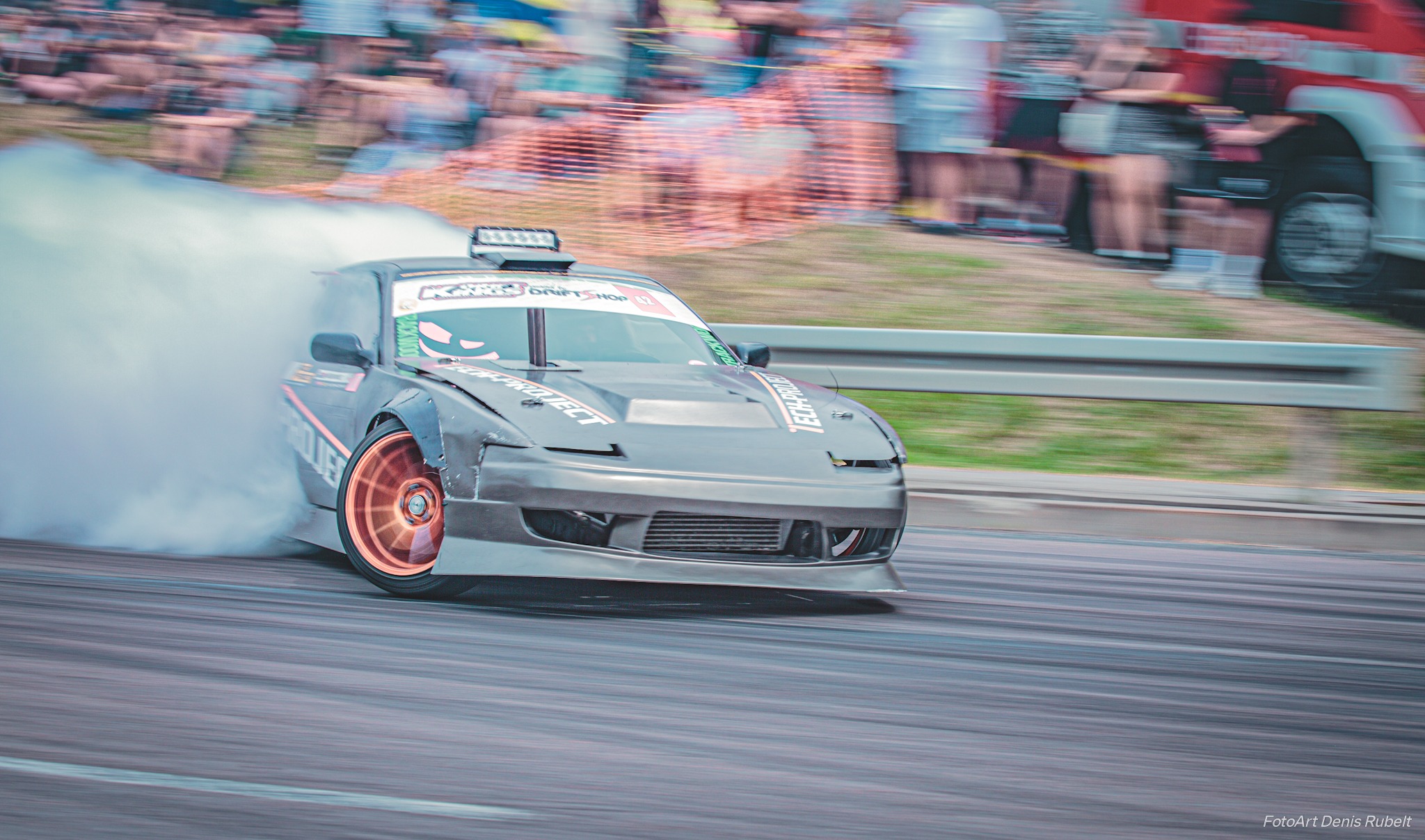 Szary samochód do driftu w trakcie jazdy, z widocznymi śladami opon i dymem, na tle rozmytej publiczności i wozu strażackiego.