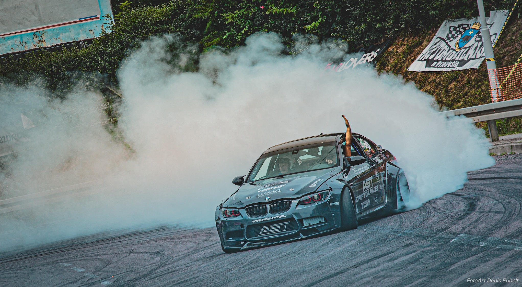 Czarny samochód BMW driftuje na zakręcie, unosząc chmurę dymu z opon, z ręką wystającą z okna, w tle zielona roślinność i banery.