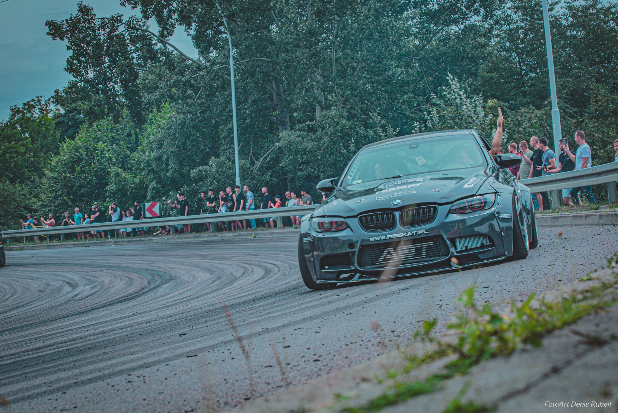 Szary samochód BMW M3 driftuje na zakręcie toru, widzowie obserwują za barierką, widoczne ślady opon na asfalcie.