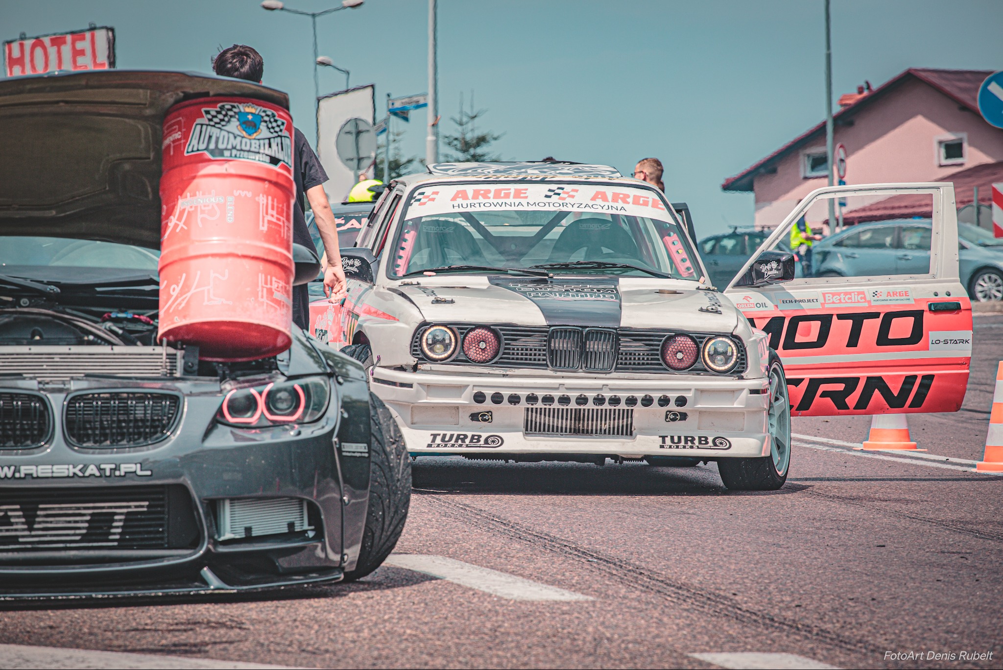 Dwa samochody wyścigowe na torze, jeden z otwartą maską i beczką z logo Automobilklubu, drugi z otwartymi drzwiami i naklejkami sponsorów