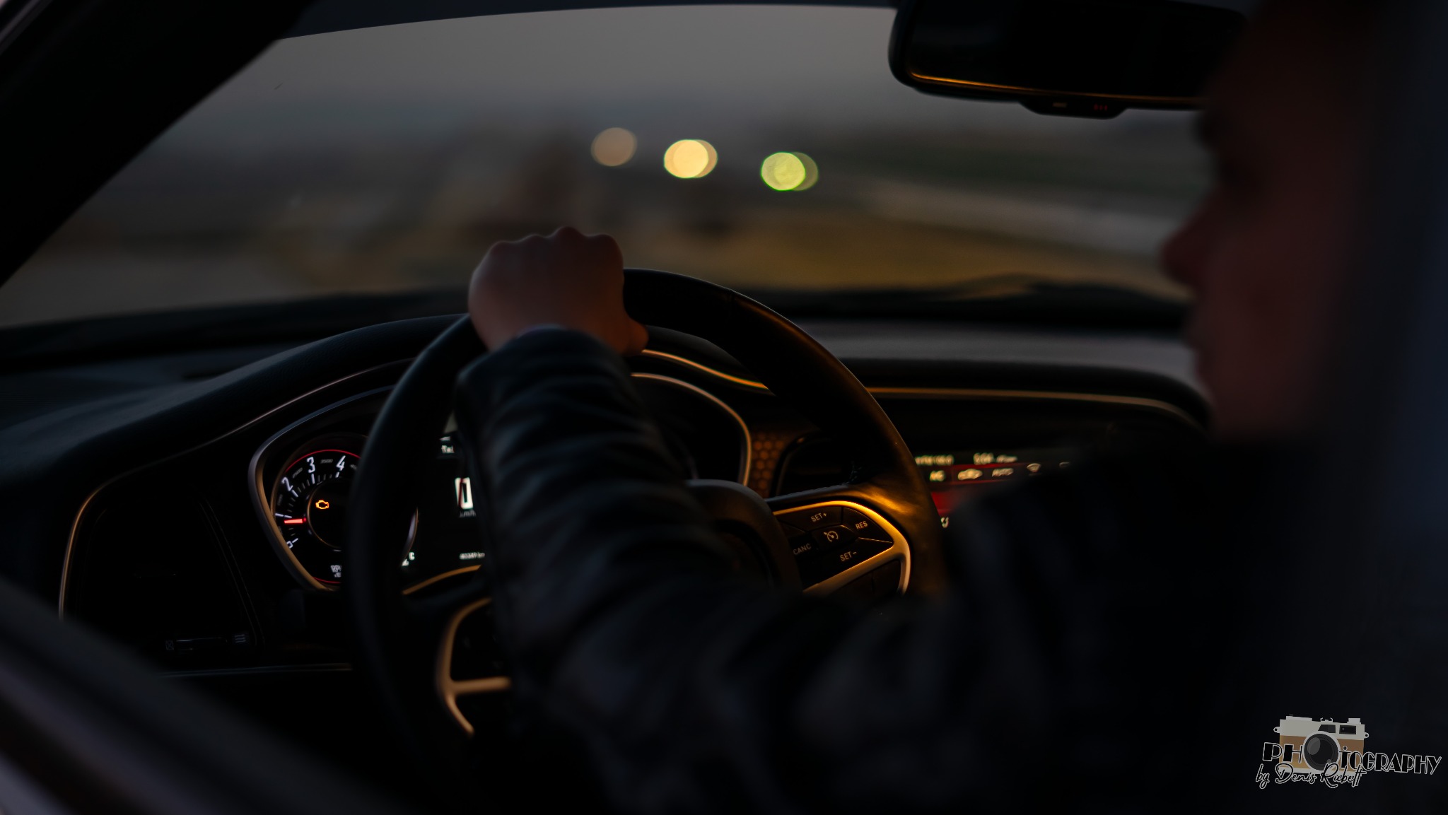 Wnętrze samochodu z perspektywy kierowcy, widoczna deska rozdzielcza, kierownica i droga w oddali z rozmytymi światłami.