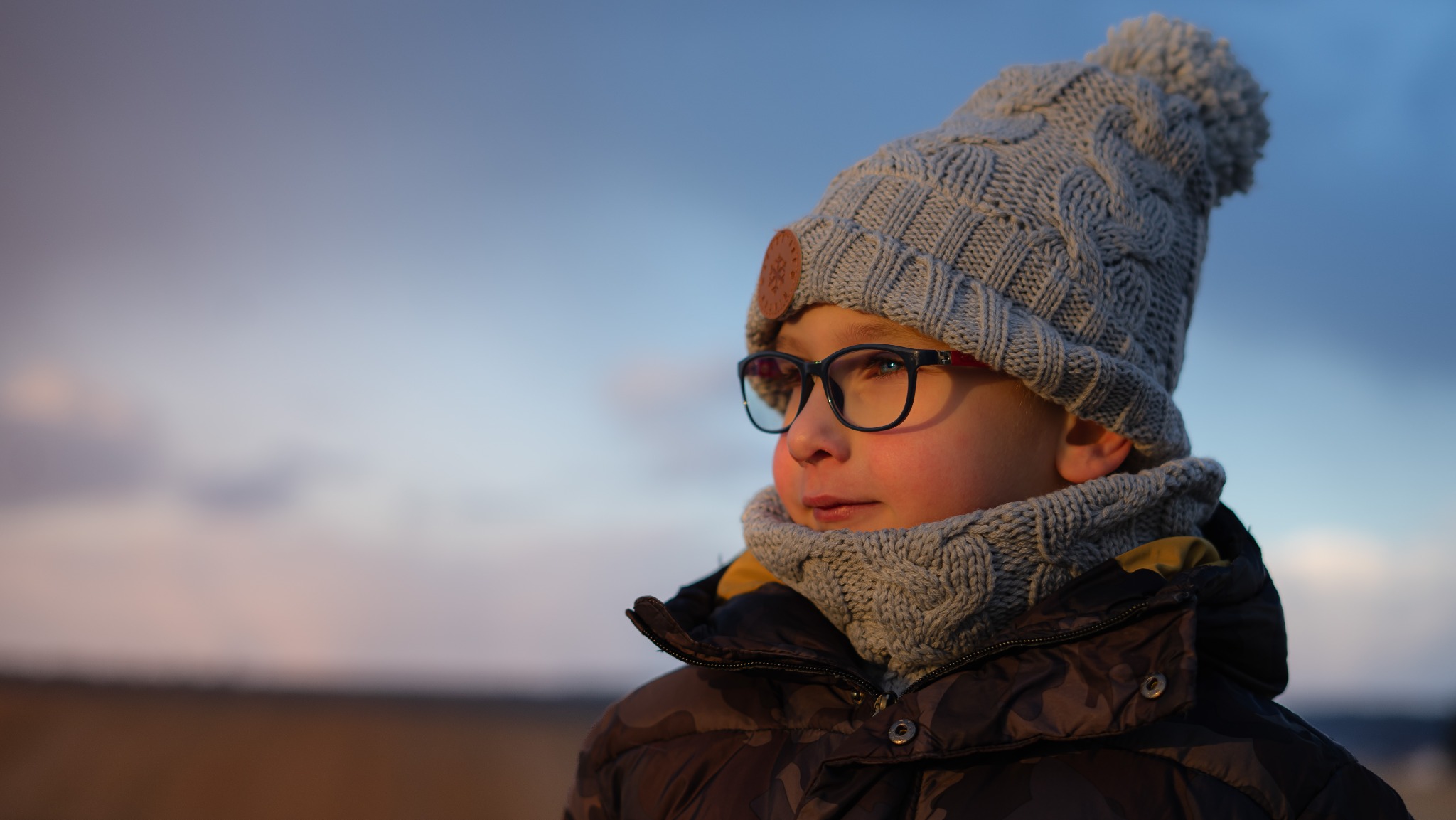 Portret chłopca w okularach i zimowej czapce z pomponem, spoglądającego w bok na tle rozmytego nieba o zachodzie słońca.