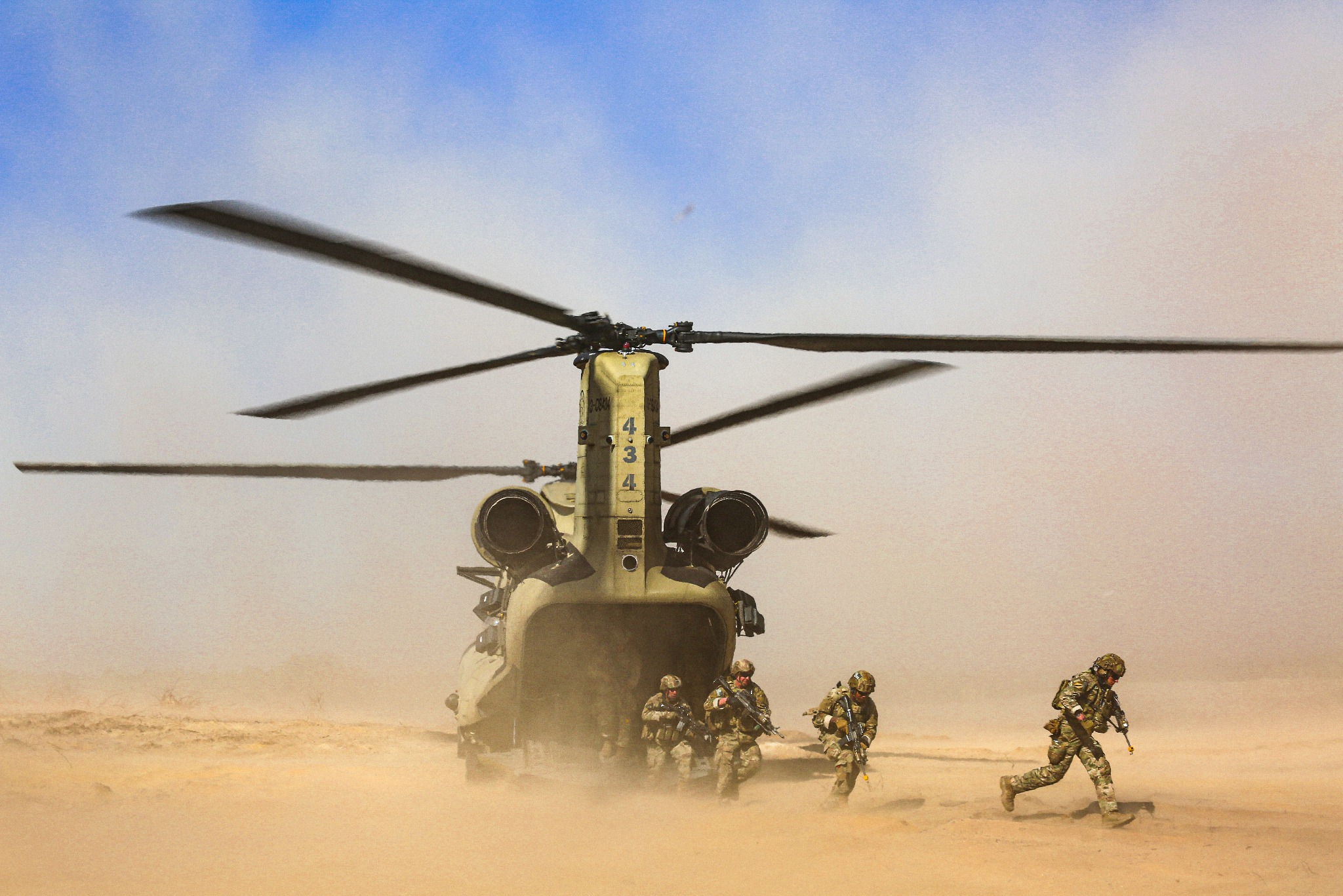 Śmigłowiec transportowy Chinook na pustyni, z którego wysiadają żołnierze w pełnym rynsztunku, unoszący się piasek.