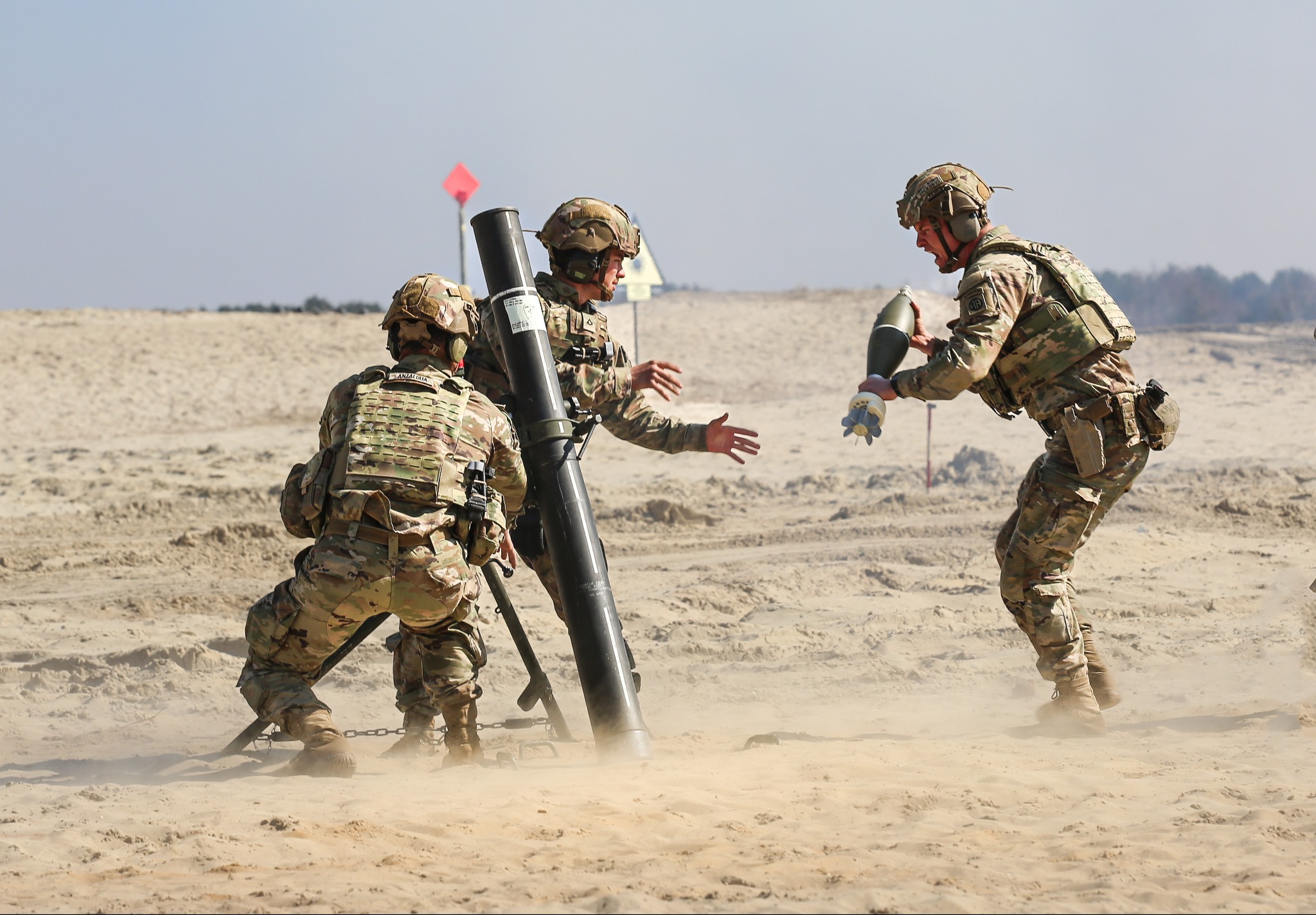 Żołnierze w kamuflażu obsługują moździerz na piaszczystym poligonie, unoszący się pył, jeden żołnierz podaje amunicję.