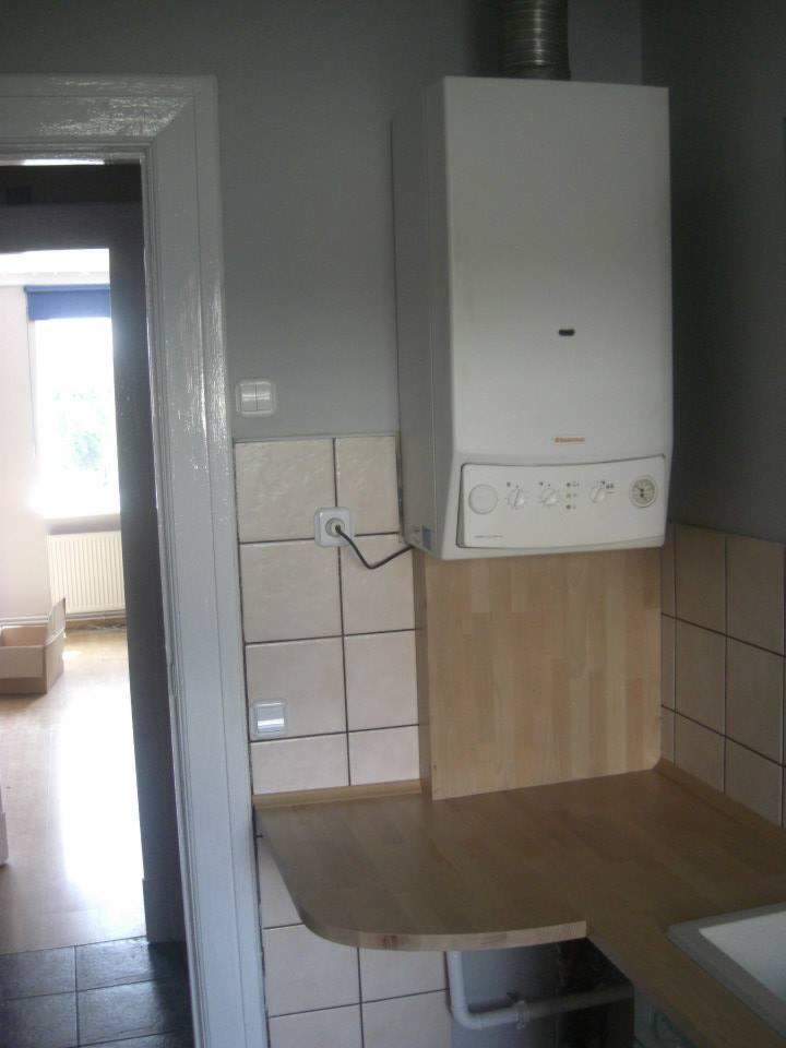 Biały piec gazowy zamontowany na ścianie nad blatem roboczym i płytkami ceramicznymi w pomieszczeniu w trakcie remontu, widoczne gniazdko elektryczne i fragment instalacji.